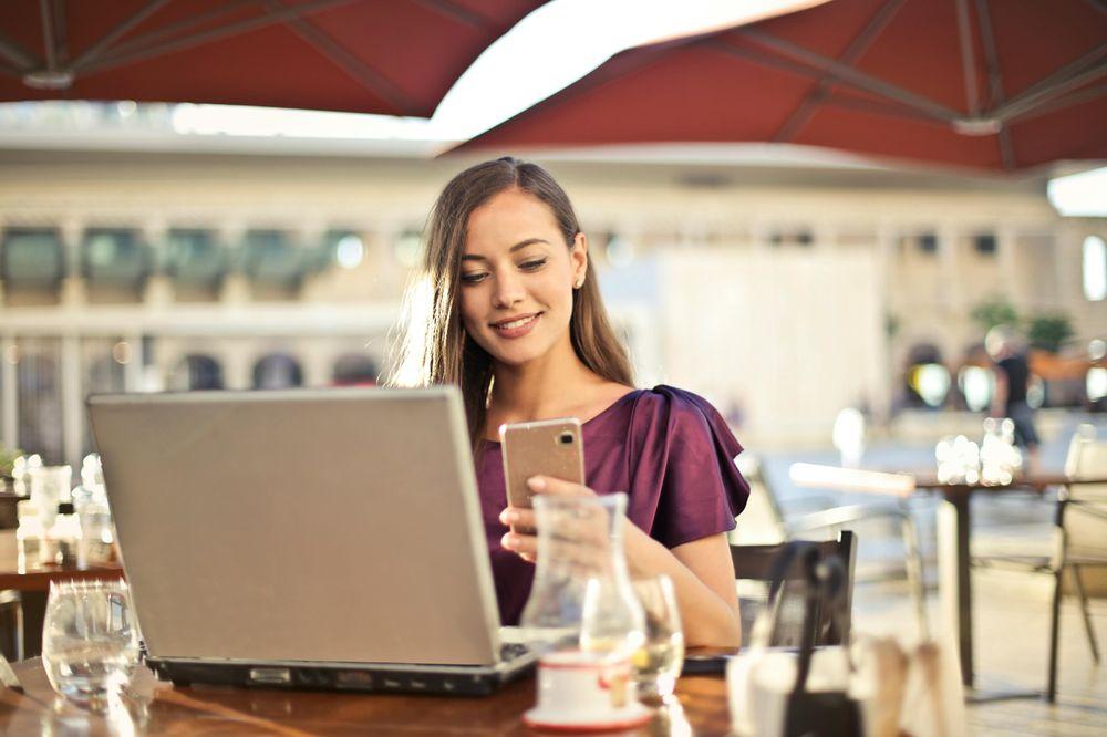 Eine Frau sitzt an einem Tisch mit ihrem Laptop vor sich und ihrem Handy in der Hand und lächelt.