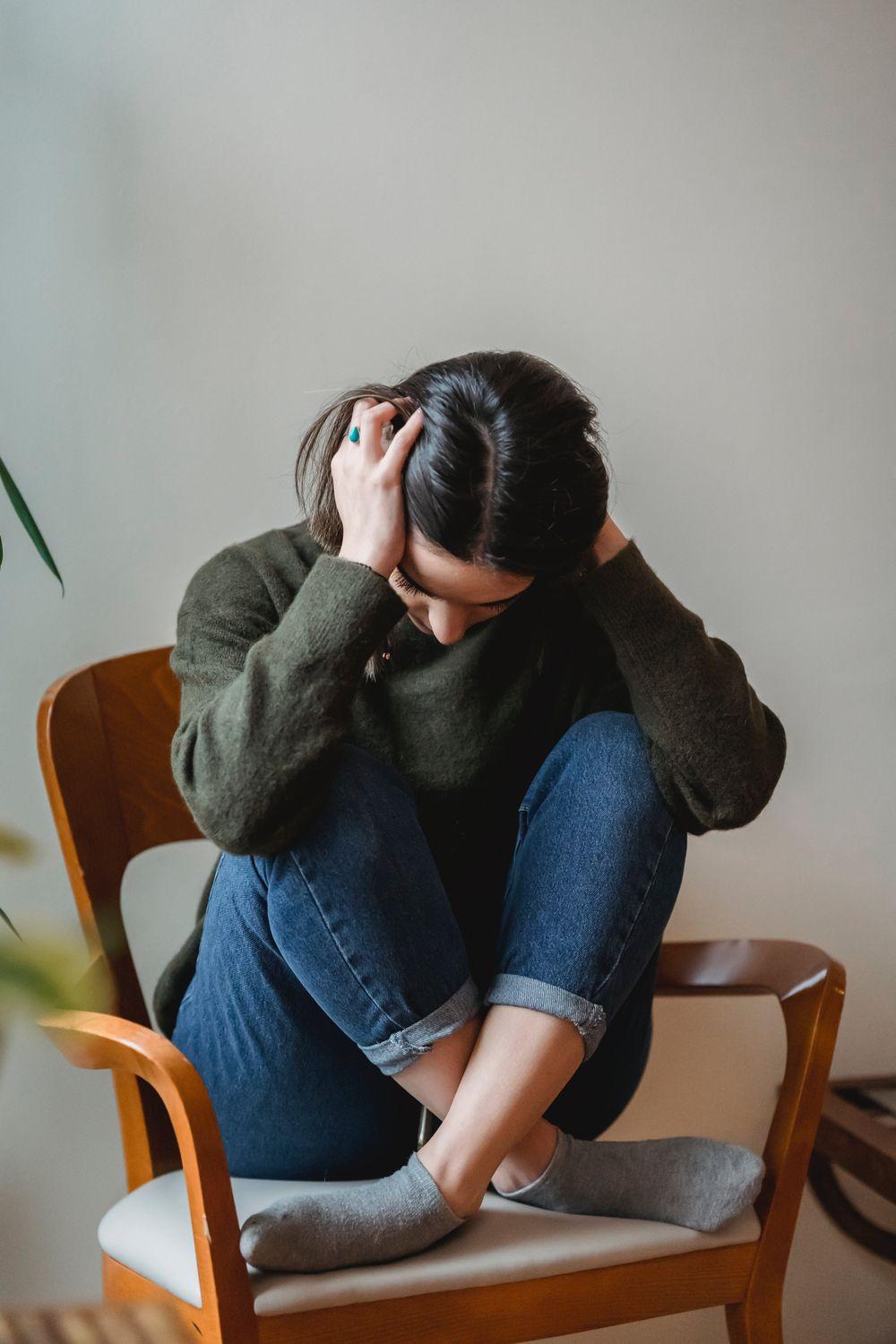 Eine Frau sitzt auf einem Stuhl, hält sich die Handflächen über die Ohren und kauert sich zusammen, weil ihre Tinnitus-Symptome psychische Probleme verursachen.