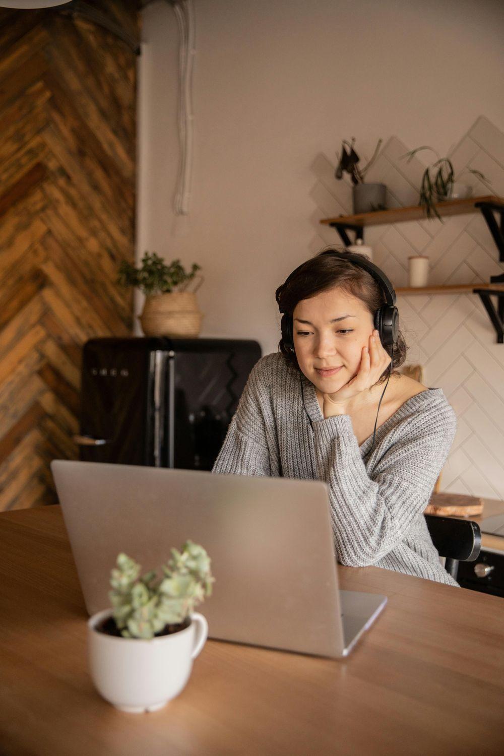Eine Frau in einem grauen Pullover sitzt zuhause in der Küche mit Kopfhörern und Laptop und macht einen kostenlosen Online-Hörtest.