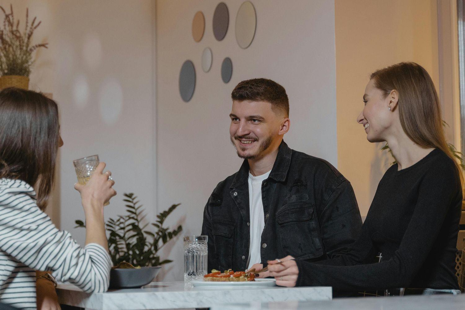Ein Mann sitzt in einer Gruppe mit zwei Frauen beim Essen und unterhält sich mit einem Lächeln.