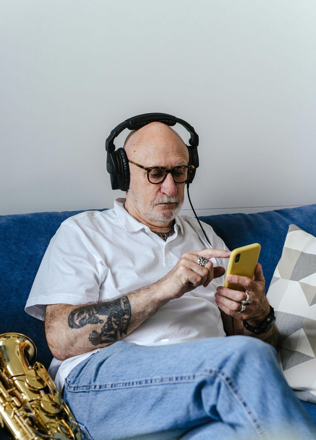 Ein älterer Mann in einem weißen T-Shirt und Kopfhörern sitzt bequem auf dem Sofa und absolviert ein Hörtraining auf seinem Smartphone.